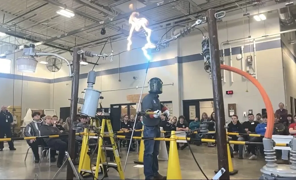 A lineman in full protective gear, including a welding helmet, performs a live electrical safety demonstration in a large indoor space. Sparks and electricity arc between high-voltage wires while an audience of students watches from their seats. Safety barriers and electrical equipment are set up for the demonstration.