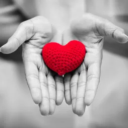 wo hands gently holding a small, crocheted red heart. The image is in black and white, with the red heart as the only element in color, creating a strong focal point.