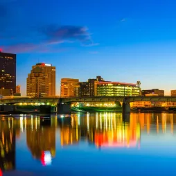 A vibrant view of the Dayton, Ohio skyline at dusk, showcasing brightly lit buildings and structures reflecting on the calm water of the river in the foreground. The skyline features a mix of modern and historic architecture, including illuminated office towers and a bridge spanning the river. Blue accent lights on tall structures near the bridge add to the colorful evening scene, with a gradient sky transitioning from deep blue to warm orange hues.