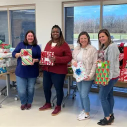 Four women stand in a room filled with holiday gifts wrapped in colorful paper, holding presents. They are smiling at the camera, dressed in festive and casual attire. Large windows behind them reveal a bright day outside, and the tables behind them are stacked with wrapped gifts for donation.