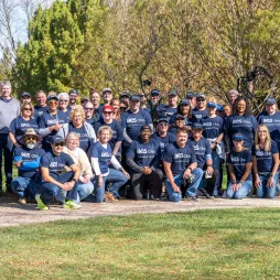 A large group of AES Ohio community volunteers poses outdoors in a park-like setting with green grass and trees. They are wearing matching navy blue shirts with the AES Ohio logo and the phrase "Your neighbor in energy." Some individuals wear sunglasses and hats, while others smile at the camera. The setting is bright and sunny, suggesting a day of community service or engagement.