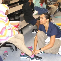 A volunteer in an AES Ohio t-shirt kneels to help a young child put on a new pair of sneakers. The child is wearing a rainbow-colored hooded jacket and beige pants. Other children and volunteers are visible in the background, participating in the shoe donation event inside a school cafeteria.