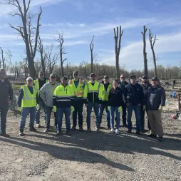 A group of AES Ohio workers in high-visibility safety vests and workwear stand together at a tornado cleanup site. The background shows a damaged landscape with barren trees, debris, and a clear blue sky. They are posing for a photo, some with hands in pockets or wearing gloves.