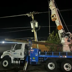 An AES Ohio utility truck with a mounted crane is parked at night, lifting a wooden power pole to repair or replace damaged electrical lines. The scene is illuminated and shows power lines and a transformer visible overhead. The truck is blue and white, featuring AES Ohio branding, and equipped for utility work. The background includes trees and a dimly lit urban setting.