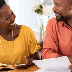 A smiling couple sits at a table in a well-lit home setting, reviewing financial documents together. The woman, wearing a yellow top, holds a pen and a smartphone, while the man, in an orange shirt, holds a piece of paper. A laptop and notebook are visible on the table, creating a collaborative and focused atmosphere. The background shows blurred home decor, adding warmth to the scene.